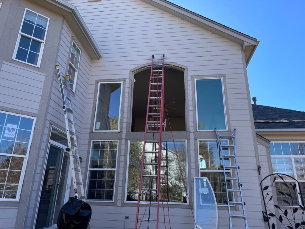view from the ground of an energy efficient window installation on a colorado home. ladder is leaning againt the siding of the home and a large decorative window is being replaced on the second story of the home